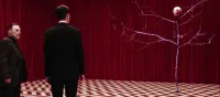 Philip Gerard and Dale Cooper look on to a tree-like creature. The floor is chevron patterned and the wall is a red curtain.