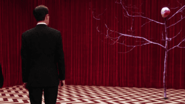 Philip Gerard and Dale Cooper look on to a tree-like creature. The floor is chevron patterned and the wall is a red curtain.