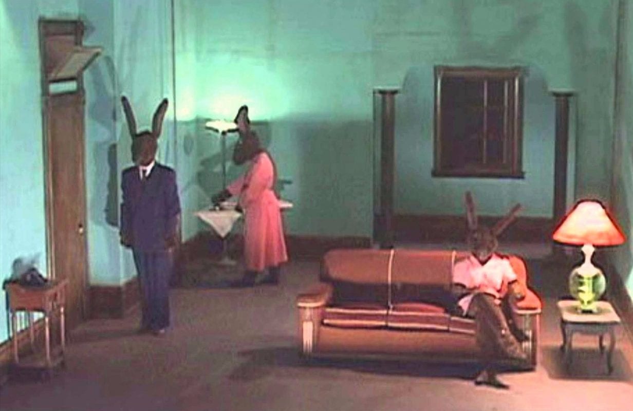 Rabbits short film by David Lynch, shows 3 rabbits in clothes in a house, one ironing, one watching tv, the other standing straight