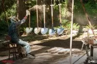 Jacoby, wearing a gasmask, spraypaints shovels that are hanging on a rack outside in the woods.
