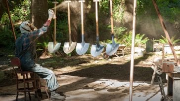Jacoby, wearing a gasmask, spraypaints shovels that are hanging on a rack outside in the woods.