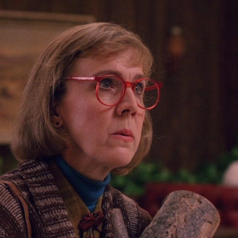 the log lady looks stern, carrying her log and wearing red rimmed glasses