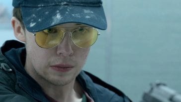 Kenny wears a hat and glasses as he holds out a gun in Black Mirror "Shut Up and Dance"