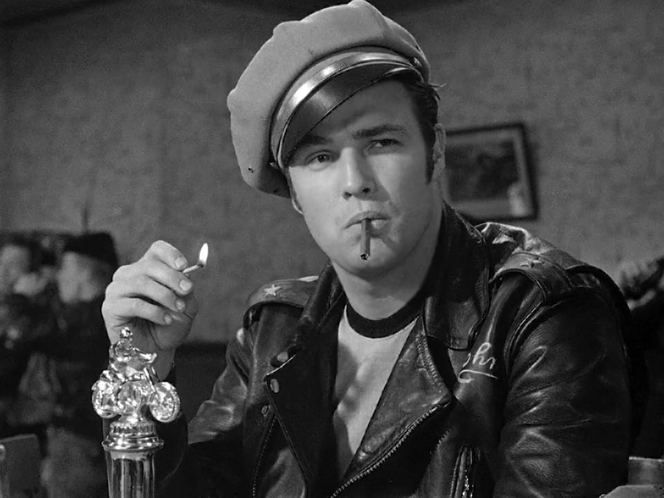 Marolon Brando wears a leather jacket with a brown hat, cigarette in his mouth and lit match in his hand.