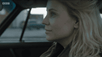 a blonde woman sits in her car staring ahead with a slight smile