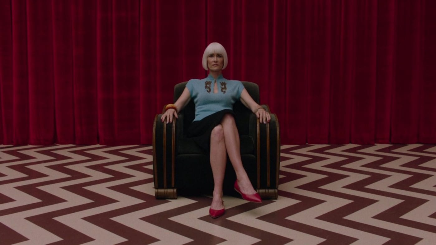 Diane Evans, sitting with her legs crossed, on a chair in the red room, chevron floor pattern and red curtains behind her.