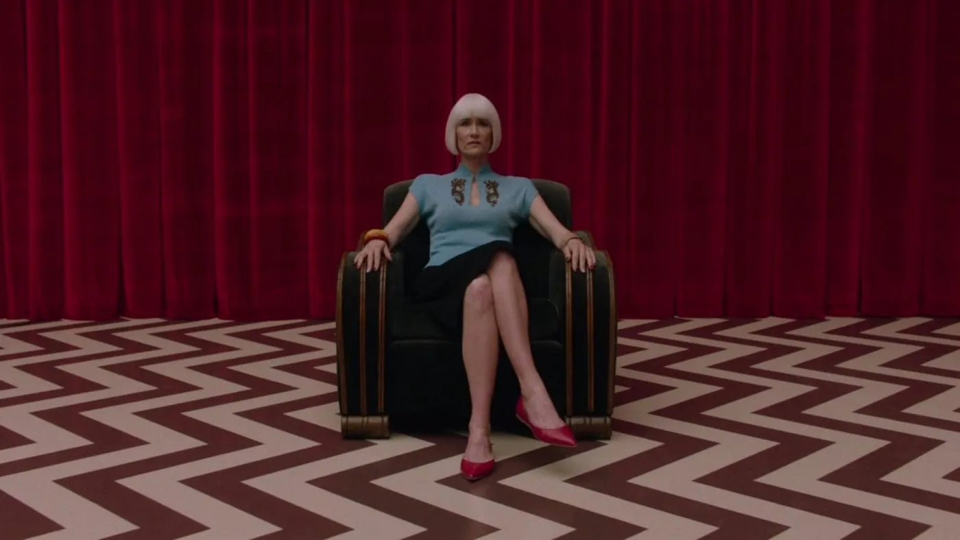 Diane Evans, sitting with her legs crossed, on a chair in the red room, chevron floor pattern and red curtains behind her.