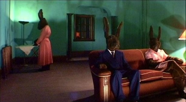 Two humanoid rabbits sit on a couch while a third irons in the background