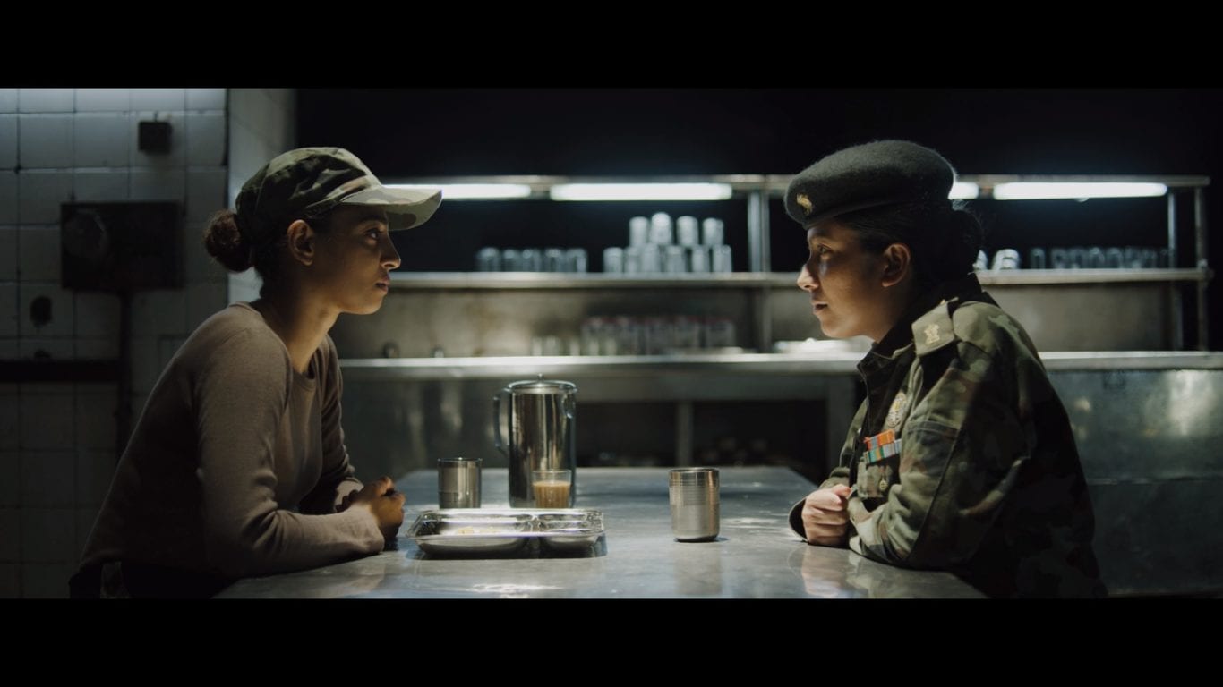 a female and male army officer sit opposite each other in a dark cafeteria kitchen