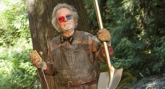 Dr Amp stands outside, holding a trigger button in one hand and a shovel in the other. He wears his trademark blue and red sunglasses.