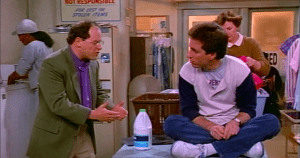 Jerry Seinfeld sits cross-legged on table while George Costanza explains something to him in the Seinfeld pilot episode