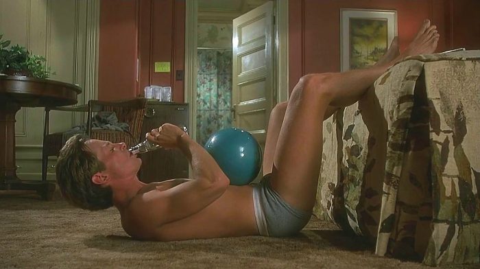 Michael J Fox in blue underwear doing sit ups with a bowling ball on his stomach and drinking alcohol from a bottle