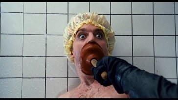 A man in a shower with a shower cap on with a mystery hand pushing a plunger over his mouth