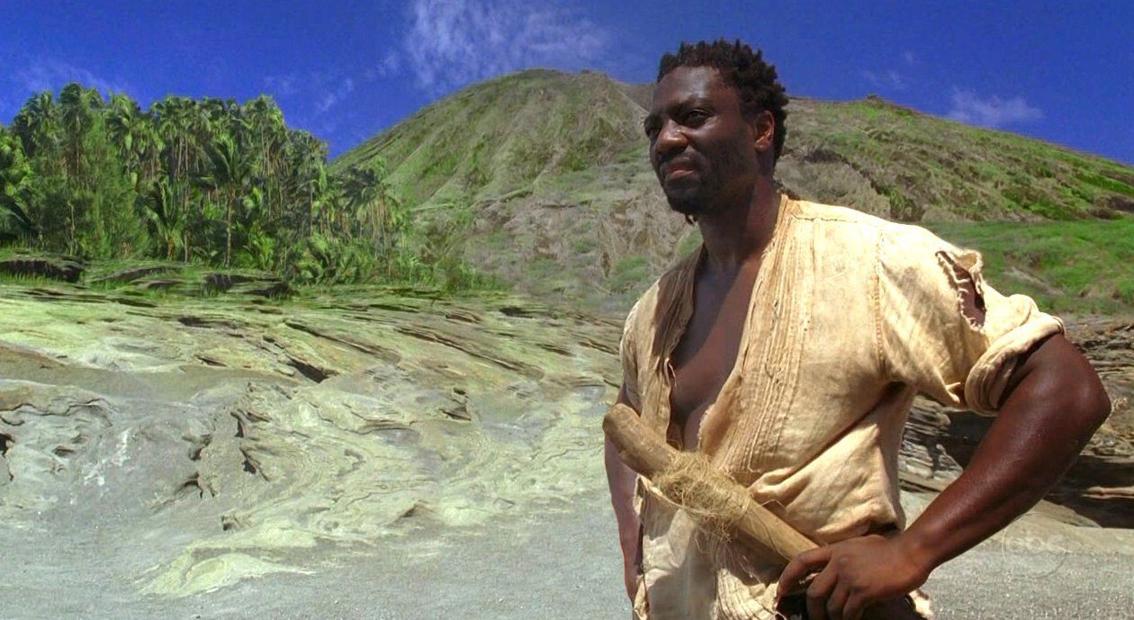 Mr. Eko stands on the beach with his hand on his hips with the mountains and forest of The Island in the distance