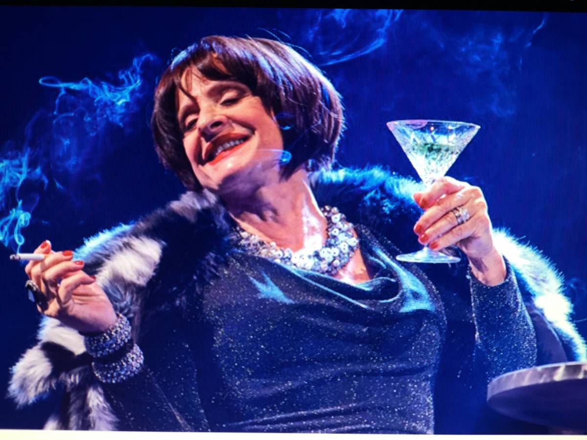 Patti Lupone as Joanne, holding a martini glass and a cigarette, looking languidly happy about something.