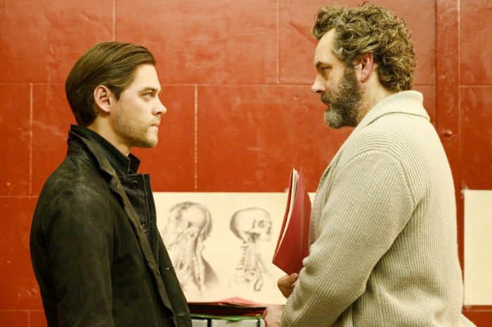 Malcolm (wearing black) and Dr. Whitly (wearing a sweater and holding a folder) stare at each other with sketches in the background