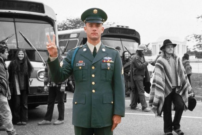 Forrest in military uniform making a peace sign with hippies in the background