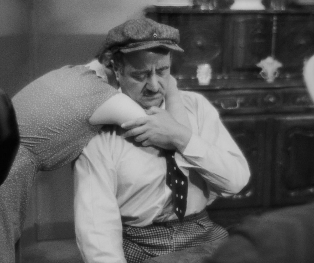 Fanny sadly hugs César around his neck as he sits on a chair.