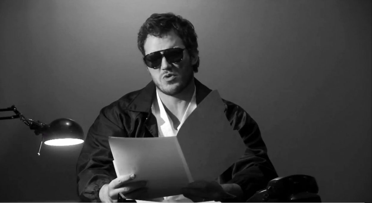 Black and white shot of Andy as Burt Macklin, FBI, sitting at a desk, wearing sunglasses and reading from a folder