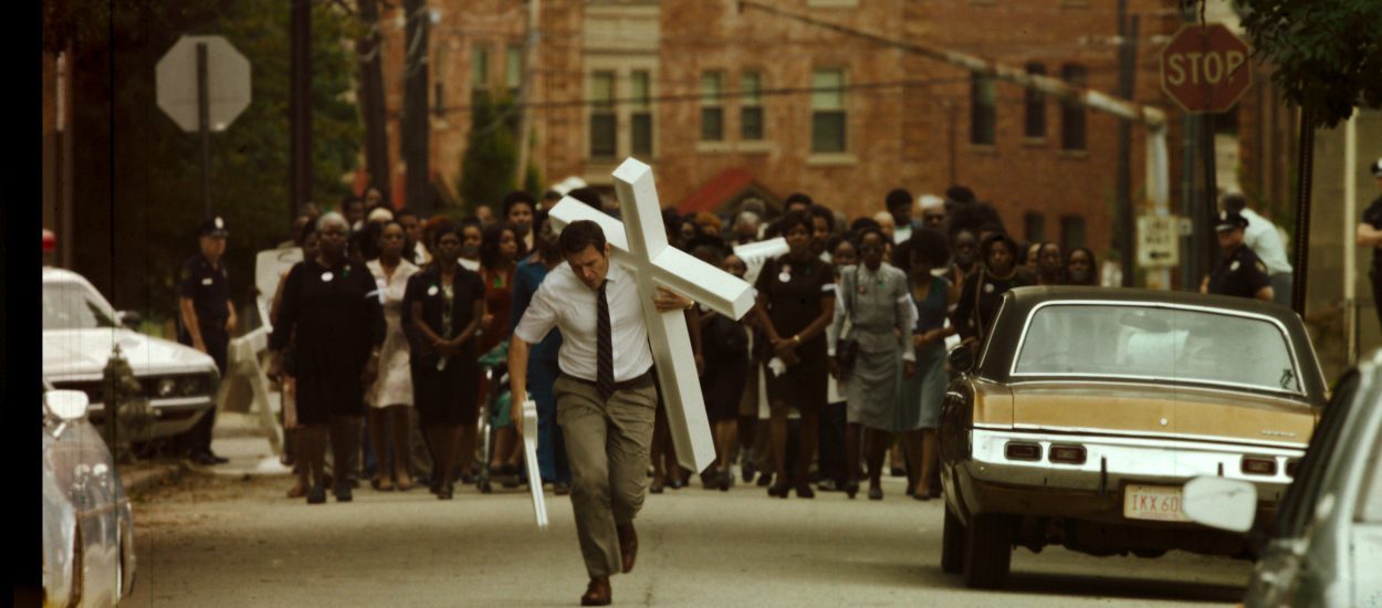 Holden walks down the streets of Atlanta carrying a cross. A large group of people are walking behind him