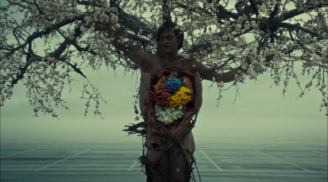 A murder victim, upright, with a tree growing out of him, and the insides of his chest and stomach cavity replaced by different flowers