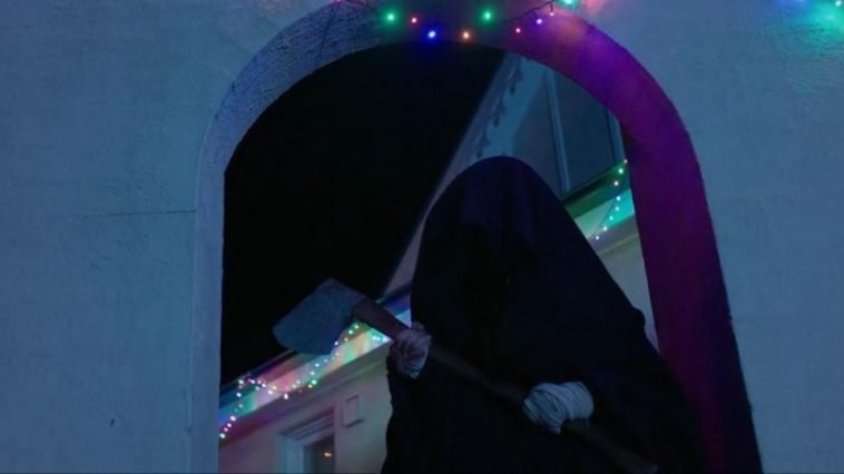 Cletus stands in a black cloak and holds an ax outside of the house in Red Christmas
