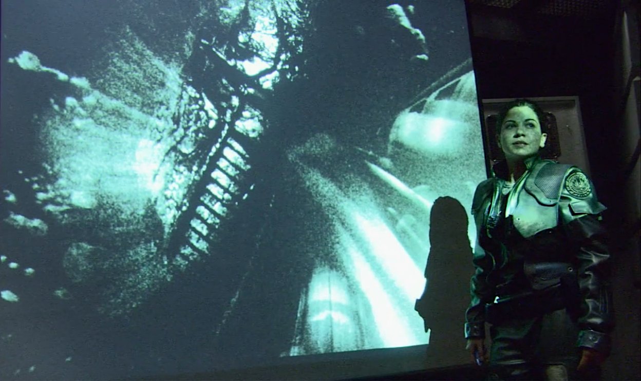 Kat stands in front of the screen showing video footage of Scar to the pilots in the briefing room