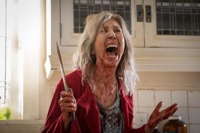 A bloodied woman standing in a kitchen screaming with a knife in her hand