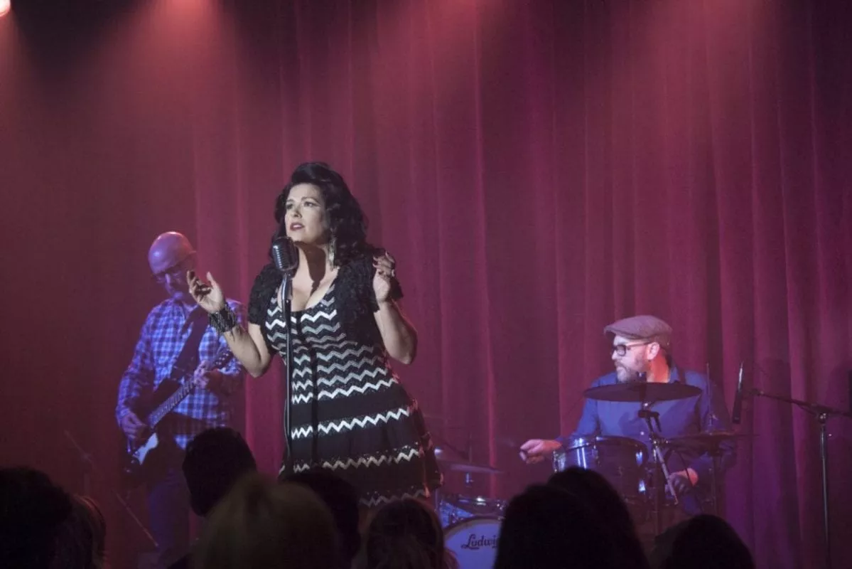 Del Rio wears a black and white chevron dress, hands outstretched. Her band mates are lit in purple, and red drapes flank them.