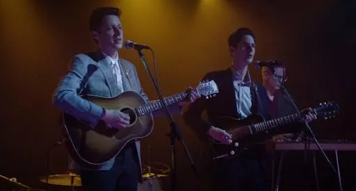 Two men sing into microphones while playing their guitars, while their background is lit in yellow.