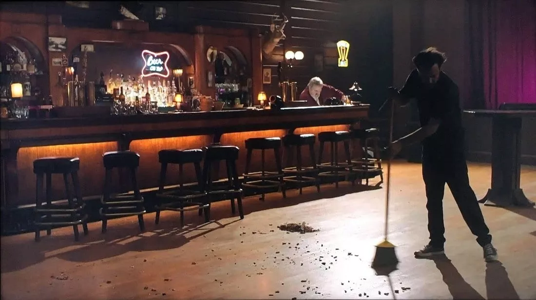 A wide shot of an empty bar with one man sweeping with a broom. Bartender is at the back cleaning the counter.