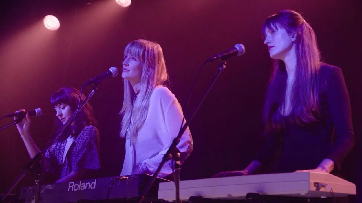 On a dutch angle, the three women sing into microphones. The woman closest to us is behind a keyboard.