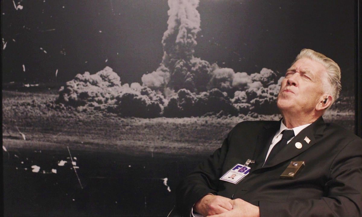 Gordon Cole leans back in his office chair whistling in front of a picture of the Los Alamos A-bomb test