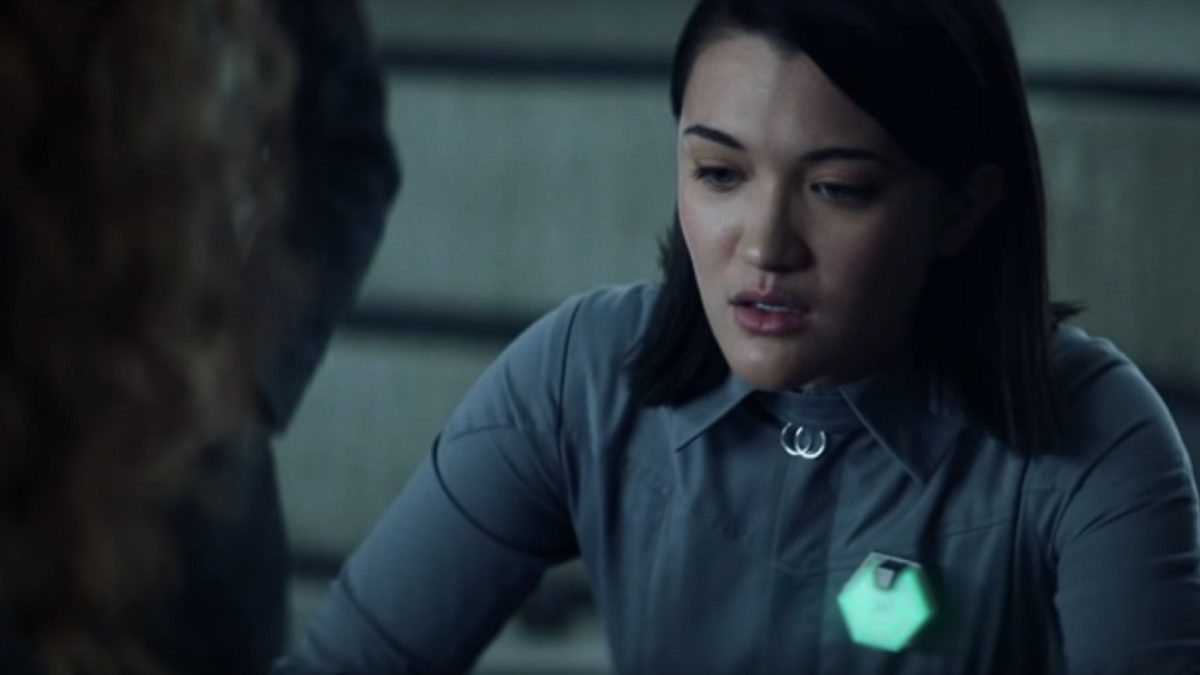 Picard S1E3 - Soji sits at a table, a hexagonal badge on her uniform is glowing green