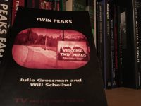 The book face out against a bookshelf background. The cover is black, with text in white, and sepiatone image of the Twin Peaks road and population sign on the side.