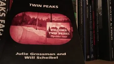 The book face out against a bookshelf background. The cover is black, with text in white, and sepiatone image of the Twin Peaks road and population sign on the side.