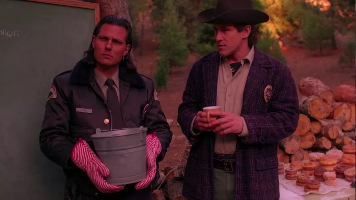 Deputy Hawk holds a pot while wearing oven mitts and Sheriff Truman holds a cup of coffee while standing next to a chalkboard in the forest.