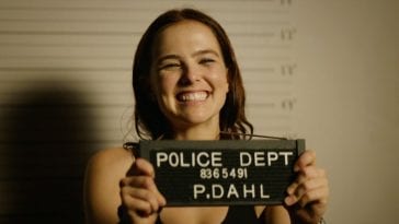 Zoey Dutch as Peg holds up a police sign and grins while getting her mugshot taken