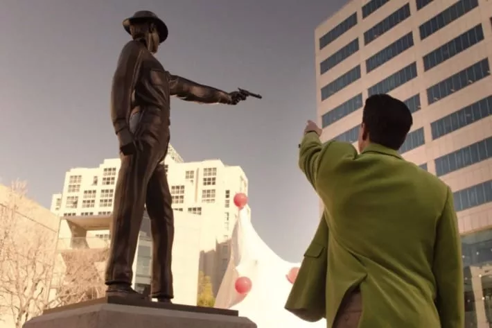 Cooper Dougie from below points his arm at a statue of a lawman holding a gun.