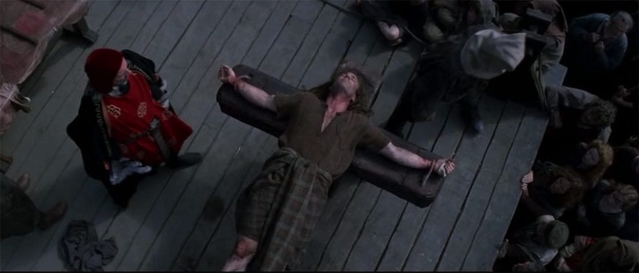 A man in red robes stands alongside William Wallace as he lies bound to a wooden crucifix