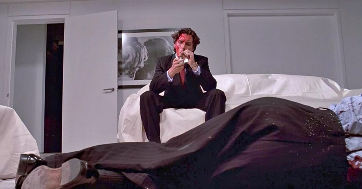 Bateman lights up a cigar whilst sitting on his sofa with blood all over his face and a corpse lying on the floor.