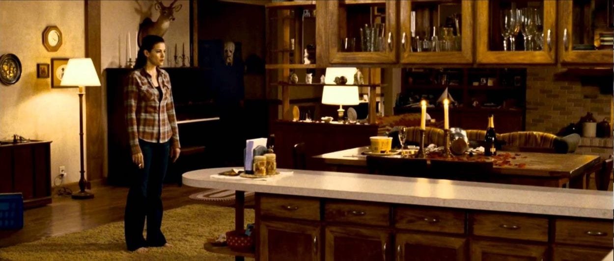 Kristin stands in the kitchen as one of the strangers watches her from the shadows