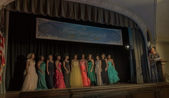 The contestants of the Miss Juneteenth pageant stand on stage underneath a large banner.