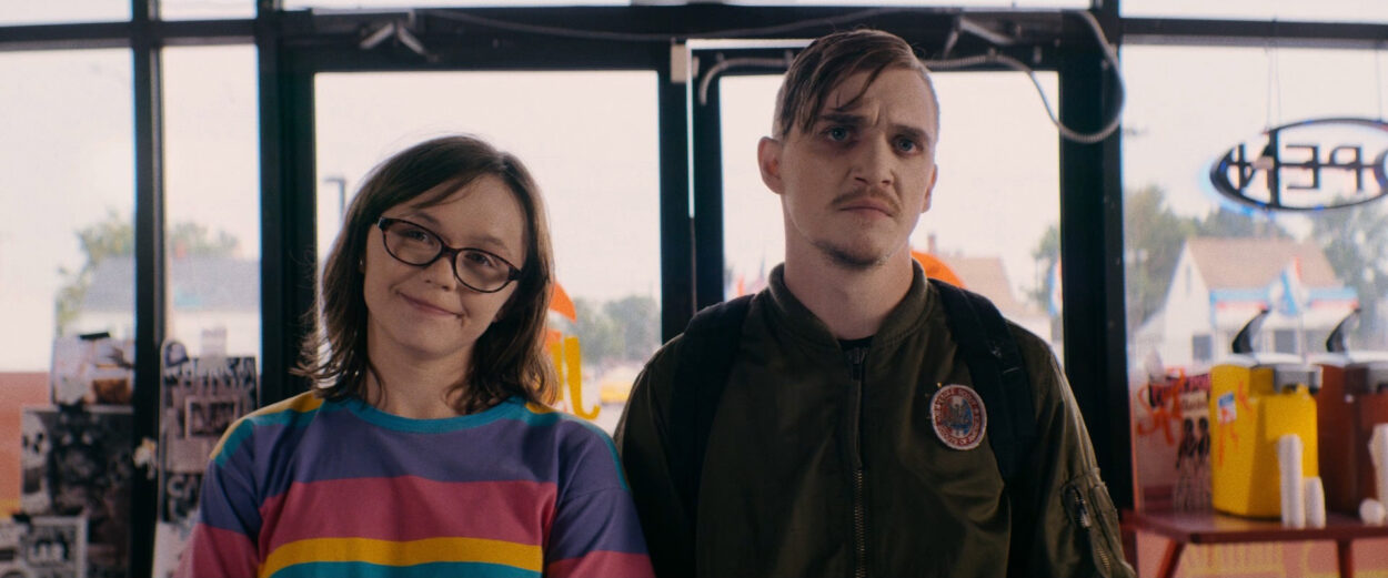 Patty (Emily Skeggs) and Simon (Kyle Gallner) standing in a fast food joint. Patty is smiling while Simon scowls.