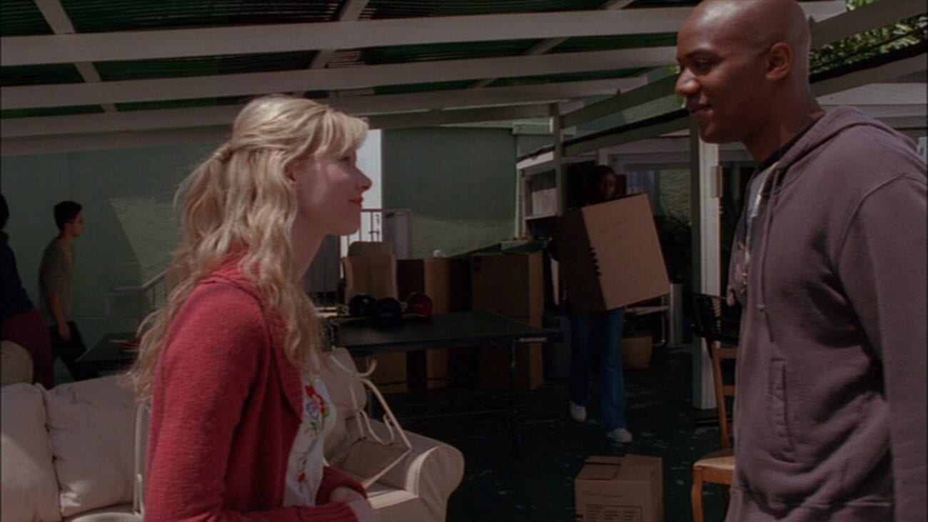 Anne and Gunn smile at each other, standing in front of Anne's shelter where people are moving boxes and furniture