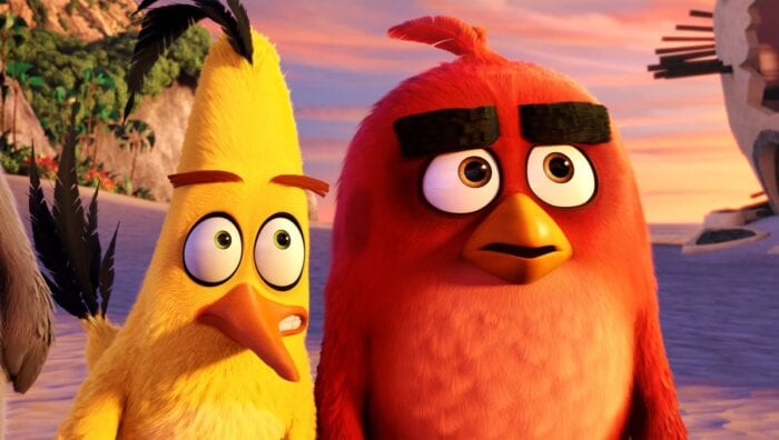 Angry Birds characters Red (right) and Chuck (left) standing on the beach outside Red's severely damaged home. Red is looking ahead with a troubled expression while Chuck leans towards him, eyes directed toward the same thing but whispering something to him through gritted teeth.