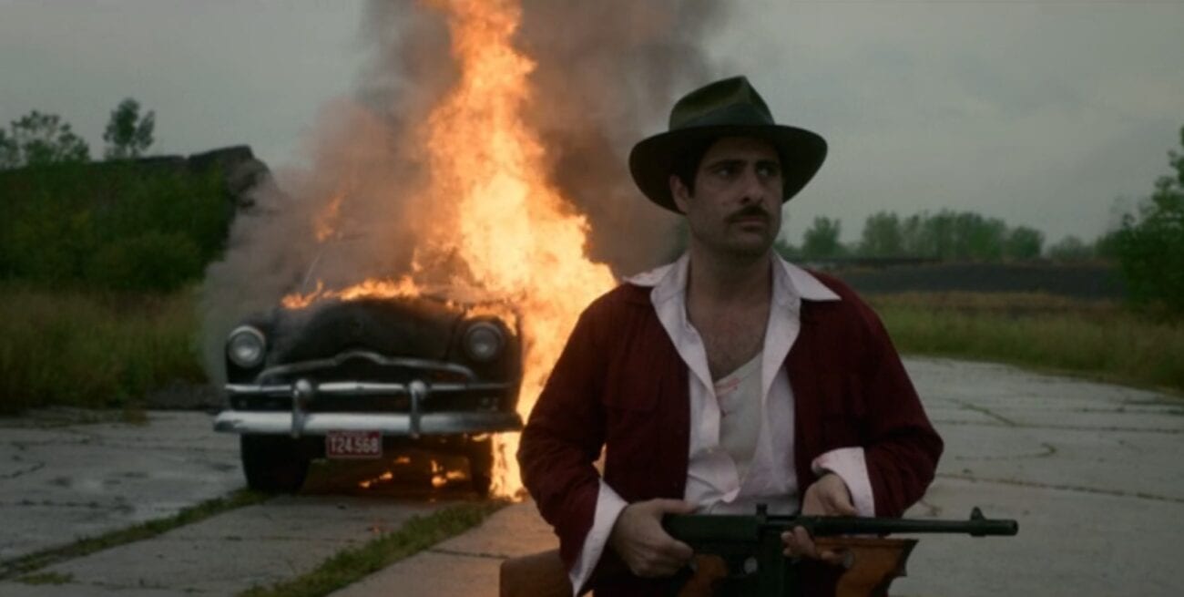 A man in a hat with a gun walks away from a burning car