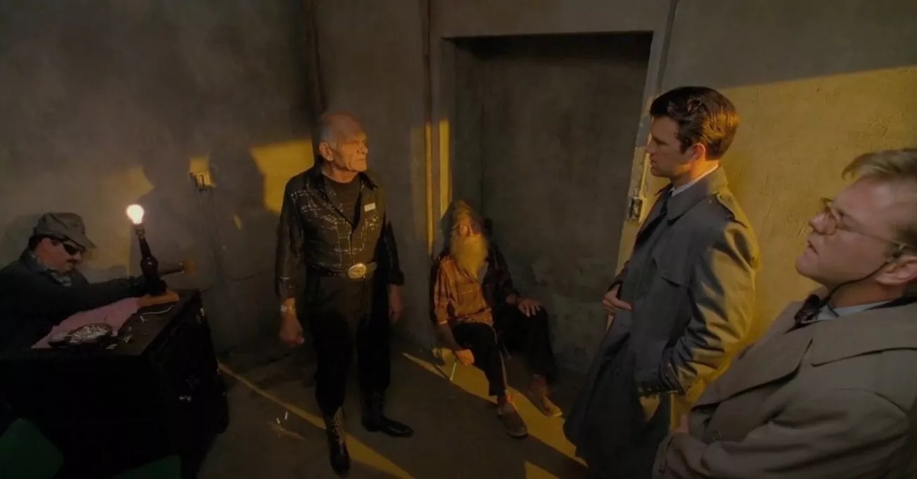 FBI agents Desmond and Stanley, in grey trench coats, question an old man dressed in black, while a man with a beard sits on the floor and another man works on a faulty lamp. 