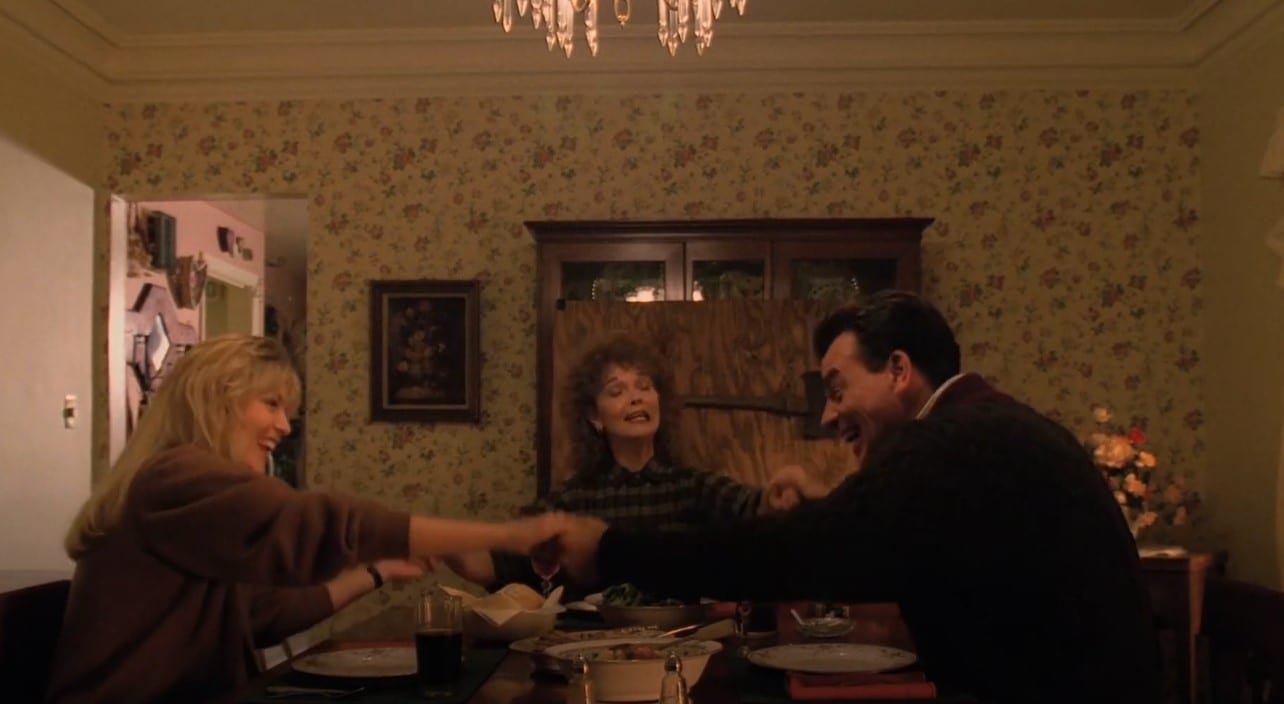 The Palmer family hold hands and laugh around the dinner table. An axe mounted on a plank of plywood is resting against the cabinet in the background. 