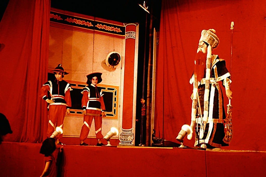Three actors stand on stage in a Chinese Opera. The background and floor are red and the two off to the left are dressed as guards while the actor to the right is dressed in full ceremonial robes of the Emperor
